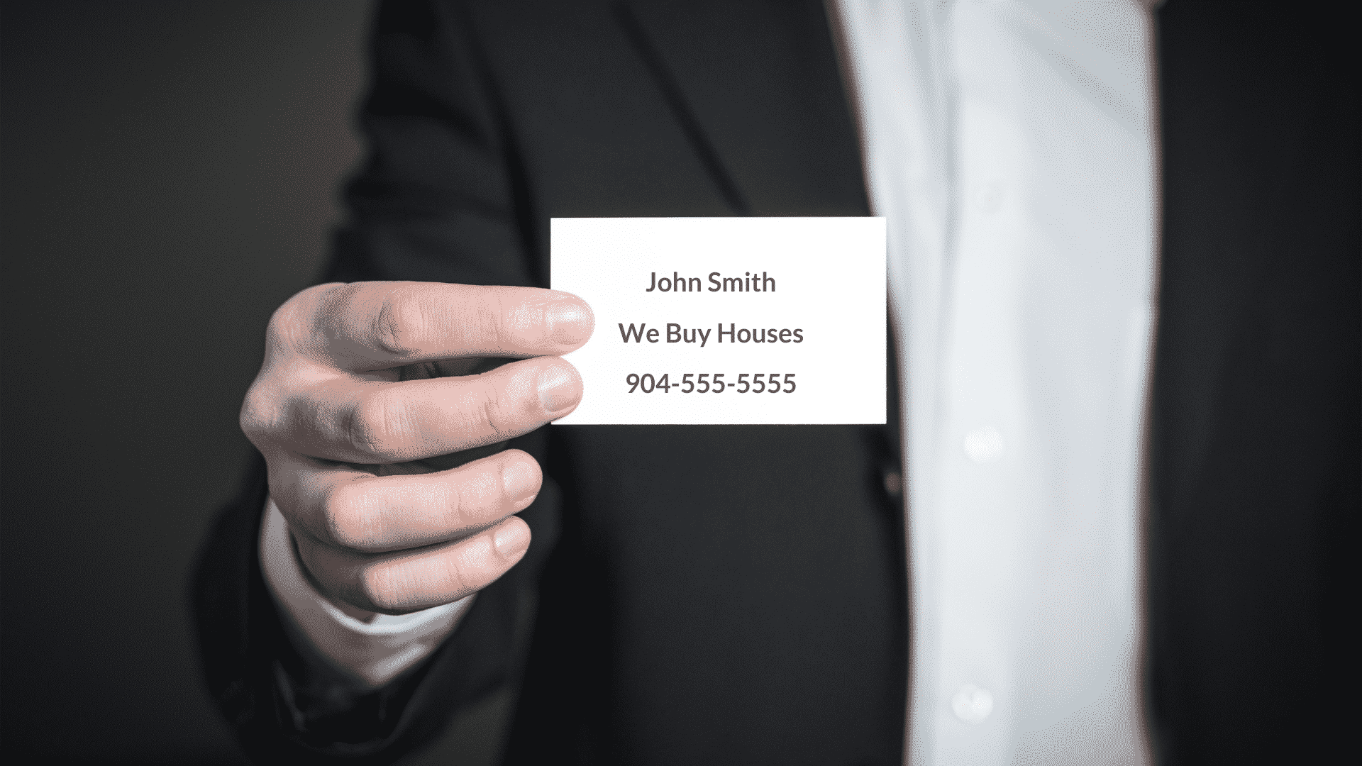 A man in a black suite holds a business card saying we buy houses