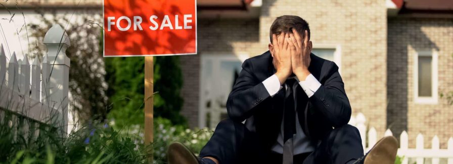 A man in a suite suite sits outside of a house with a for sale sign looking miserable. How can you move if your house hasn't sold yet in FL?