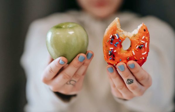 A woman holds a doughnut and an apple in her hands asking you to choose