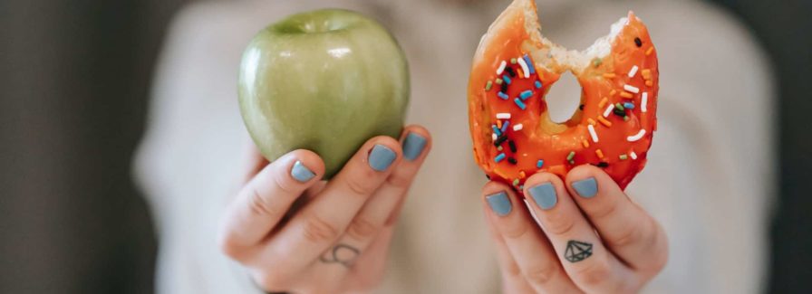 A woman holds a doughnut and an apple in her hands asking you to choose