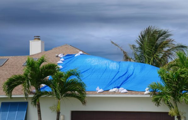 Hurricane damaged roof on house with a blue plastic tarp over hole in the shingles and rooftop.