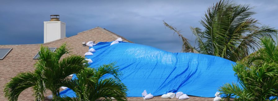 Hurricane damaged roof on house with a blue plastic tarp over hole in the shingles and rooftop.